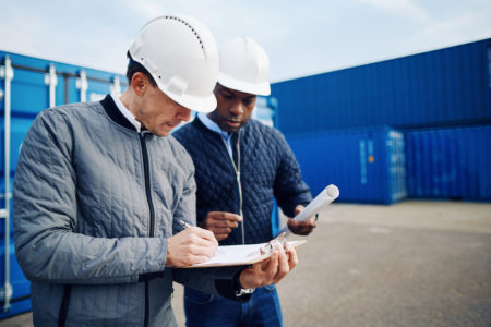 Two engineers standing by freight containers on a large commercial dock discussing shipping logistics together